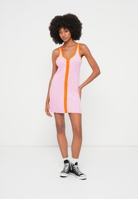 Femme à la coiffure afro portant une mini-robe rose sans manches avec des bordures orange et des baskets montantes noires, debout devant un fond blanc.