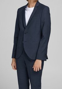 Giacca e pantaloni da abito blu navy in tessuto strutturato, con chiusura a un bottone e taschino sul petto; indossati sopra una t-shirt bianca.