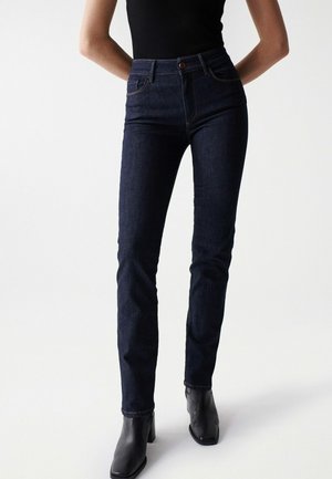 Personne portant un jean slim taille haute bleu foncé avec poches avant et des bottines noires, debout devant un fond blanc uni.