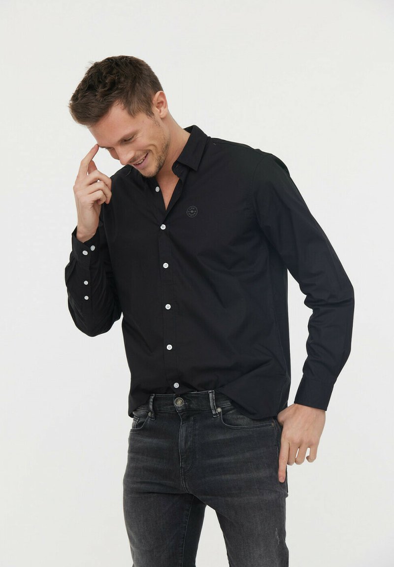 Chemise noire à manches longues avec un col à boutons, boutons blancs et un logo discret sur la poitrine, associée à un jean foncé.