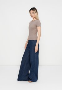 Femme se tenant de côté, portant un haut marron ajusté à manches courtes et un pantalon large bleu foncé, devant un fond blanc uni.