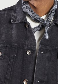 Primer plano de una persona que lleva una chaqueta de mezclilla oscura sobre una camisa blanca con un pañuelo de paisley en blanco y negro atado alrededor del cuello.