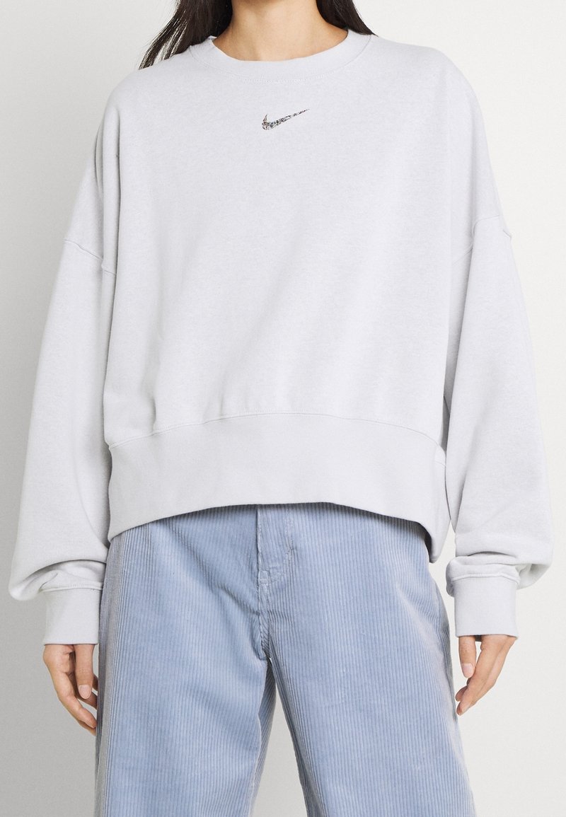 Personne portant un sweat-shirt blanc ample Nike avec un petit logo noir et un pantalon en velours côtelé bleu clair, debout devant un fond uni.