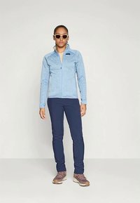 Casaco de fleece azul claro com fecho éclair à frente, combinado com calças de tom navy e ténis castanhos. Os acessórios incluem óculos de sol brancos.