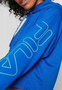 Närmre av en person som bär en ljusblå hoodie med en stor ljusblå konturerad logotyp på ärmen och märkta dragband.