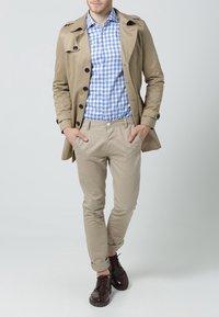 Beżowy płaszcz trench, niebieska i biała koszula w kratę, jasne khaki spodnie, brązowe buty. Model stoi z rękami w kieszeniach, neutralne tło.