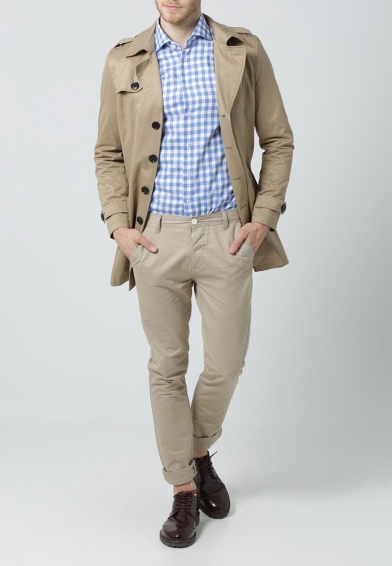 Beżowy płaszcz trench, niebieska i biała koszula w kratę, jasne khaki spodnie, brązowe buty. Model stoi z rękami w kieszeniach, neutralne tło.