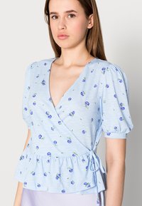 Blusa envolvente azul clara com mangas curtas e bufantes, apresentando um padrão floral. O tecido tem uma textura subtil e uma bainha peplum com um detalhe amarrado.