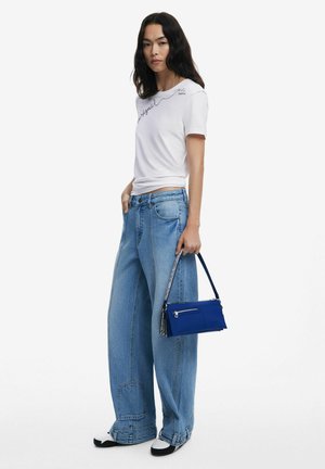 White t-shirt with black handwritten text, paired with light blue wide-leg jeans and a small blue handbag with silver accents.