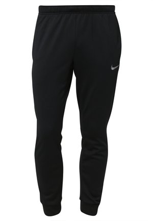 Mann trägt schwarzes Nike-T-Shirt und Jogginghose mit weißen und neongrünen Nike-Sneakers, steht mit den Händen in den Taschen.