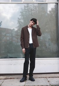 Brūna pogu krekls virs baltas T-krekla, apvienots ar melniem biksēm un melnām kurpēm. Gluda auduma ar redzamām pogām un apkakles detaļām.