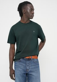 T-shirt en coton vert foncé avec un col rond et des manches courtes, associé à un jean en denim bleu et une ceinture en cuir marron.