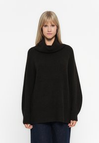 Oversized schwarzer Strickpullover mit einem Schalkragen, gerippter Textur und weiten Ärmeln, kombiniert mit dunkelblauen weit geschnittenen Jeans.