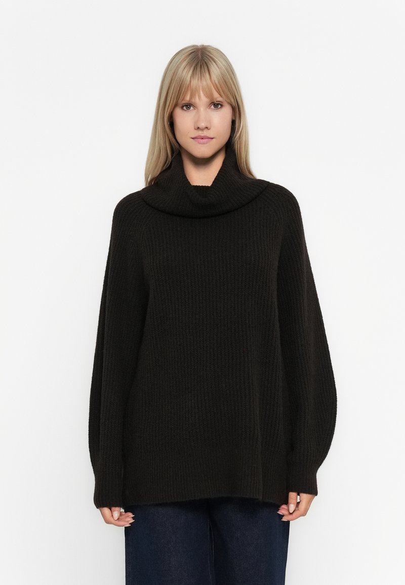 Oversized schwarzer Strickpullover mit einem Schalkragen, gerippter Textur und weiten Ärmeln, kombiniert mit dunkelblauen weit geschnittenen Jeans.