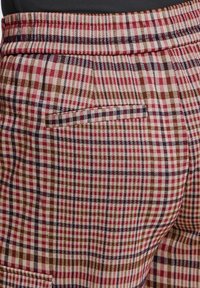 Gros plan de pantalons à carreaux rouges, marron et blancs avec une poche arrière horizontale et un détail visible à la ceinture.
