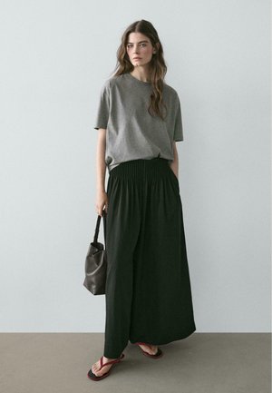 Frau mit langen braunen Haaren, die ein graues kurzärmeliges Shirt, schwarze plissierte Hosen mit weitem Bein, rote Sandalen trägt und eine schwarze Handtasche hält.