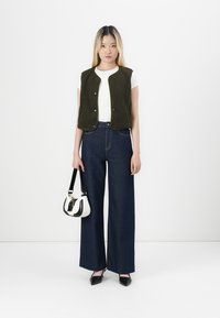 Mørkegrønn fleecevest over en hvit skjorte, kombinert med høyt liv mørk denim bukser med vide ben. Tilbehøret inkluderer en liten svart og hvit veske.