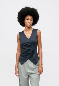 Mulher com cabelo afro vestindo uma blusa sem mangas escura e calças cinza claro com riscas, de pé contra um fundo claro e liso.