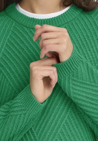 Pulover verde tricotat cu manșete ribbate, având un model texturat. Mâinile sunt așezate aproape de guler, evidențiind moliciunea materialului.