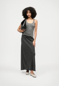 Ženska s kodrastimi lasmi, oblečena v sivo majico brez rokavov in dolgo črno krilo, drži veliko črno torbico, postavljena ob enobarvni ozadju.