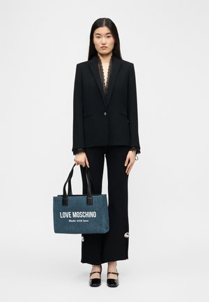 Γυναίκα με μαύρο σακάκι και παντελόνι κρατώντας μπλε τσάντα tote Love Moschino με μαύρες λαβές, στέκεται μπροστά σε λευκό απλό φόντο.