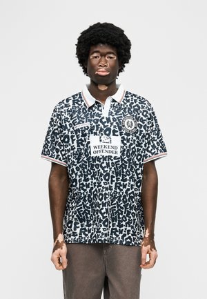 Giovane uomo con vitiligine che indossa una polo a motivi bianco e nero con la scritta "Weekend Offender" e pantaloni marroni casual, in piedi davanti a uno sfondo bianco.