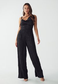 Black jumpsuit with a textured, crinkled design. Features a cowl neckline and wide-leg cut, paired with strappy sandals.