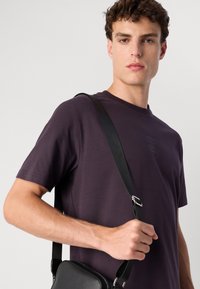 Fiatal férfi sötét lila pólót visel, és egy fekete keresztpántos táska pántját fogja egy egyszínű világos háttér előtt.