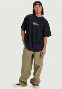 Oversized black cotton t-shirt with red graphic logo, paired with loose khaki pants and black and white sneakers. Casual streetwear look.