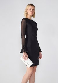 Μαύρο φόρεμα με διαφανείς, φλαμπέ μανίκες και τονική λεπτομέρεια. Το μοντέλο κρατάει μια λευκή τσάντα φάκελο με ασημένια λεπτομέρεια, σε απλό φόντο.