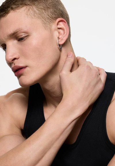 Jeune homme aux cheveux courts portant un débardeur noir, touchant son cou d'une main et portant une boucle d'oreille argentée en forme d'anneau à l'oreille gauche.