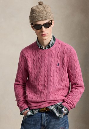 Jeune homme portant un bonnet en maille beige, des lunettes de soleil foncées, un pull torsadé rose, une chemise à carreaux verte et un jean bleu, les mains dans les poches.