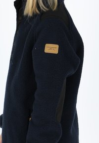 Giacca in pile blu marino con accenti neri, caratterizzata da una superficie testurizzata, un'etichetta sul petto e una tasca laterale con zip.
