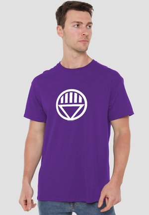 Mann trägt ein lila T-Shirt mit einem weißen runden Emblem, das fünf vertikale Balken über einem weißen Dreieck zeigt, stehend vor einem schlichten Hintergrund.
