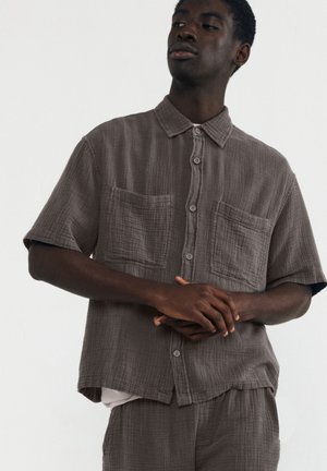 Camicia grigia a maniche corte con bottoni, realizzata in un tessuto testurizzato, con due tasche frontali e una vestibilità ampia. Indossata con una canottiera bianca.