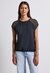 Zwarte top met korte mouwen, doorzichtige stofaccenten op de schouders, comfortabele pasvorm, ronde halslijn en een gestructureerde afwerking; gecombineerd met lichtblauwe jeans.