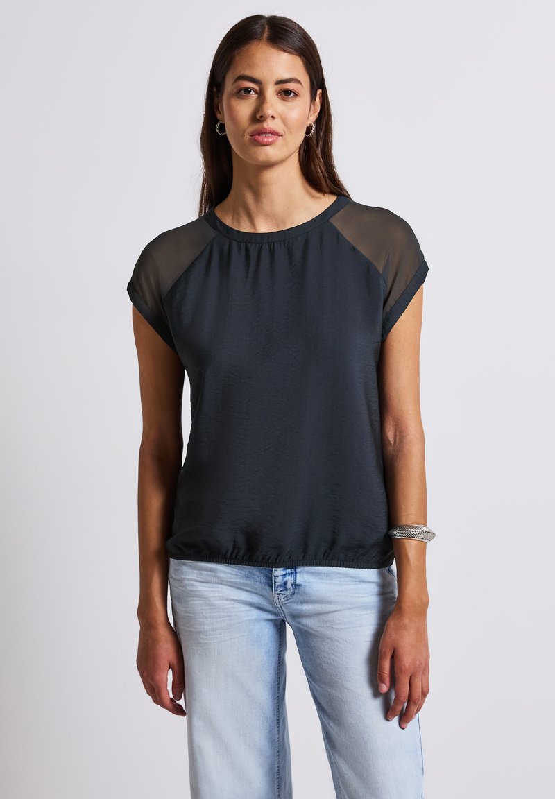 Zwarte top met korte mouwen, doorzichtige stofaccenten op de schouders, comfortabele pasvorm, ronde halslijn en een gestructureerde afwerking; gecombineerd met lichtblauwe jeans.