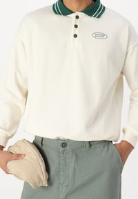 Kremowy sweter z zielonym, prążkowanym kołnierzem i trzema ciemnozielonymi guzikami. Z przodu znajduje się logo "Kaotiko Society". Ręka trzyma beżowy kapelusz.