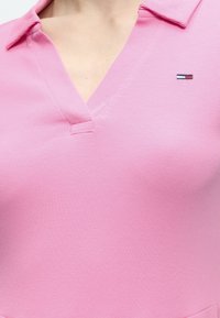 Κοντινό πλάνο ενός ατόμου που φοράει ένα ροζ ριμπ πικέ πόλο με ανοιχτό γιακά και μικρό τετράγωνο λογότυπο κόκκινου, λευκού και μπλε στο στήθος.