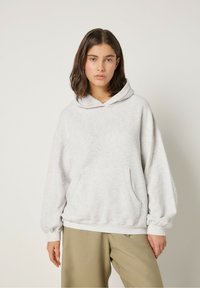 Sudadera de gran tamaño en color gris claro, fabricada en un tejido suave, que cuenta con un bolsillo delantero, capucha con cordón y puños elásticos, combinada con pantalones beige.