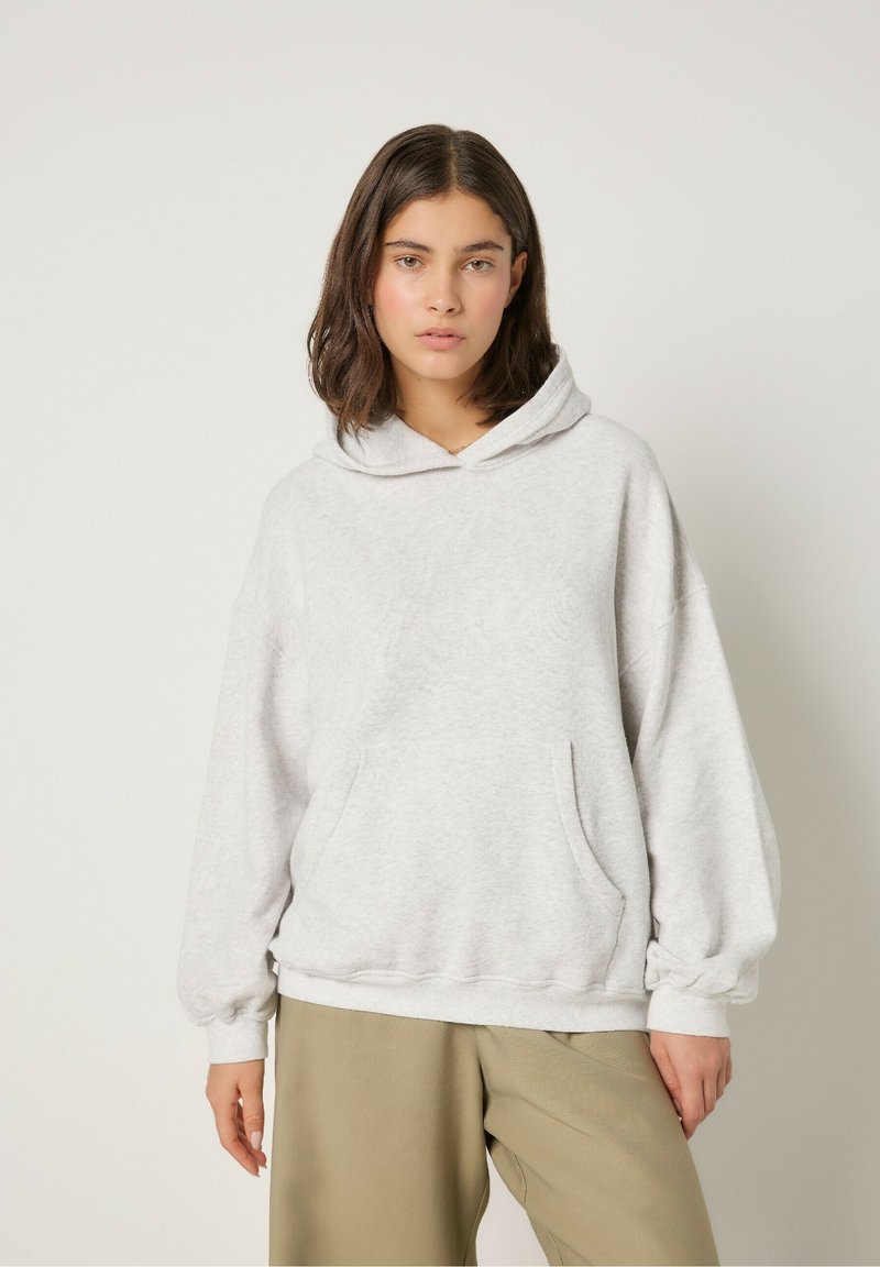 Sudadera de gran tamaño en color gris claro, fabricada en un tejido suave, que cuenta con un bolsillo delantero, capucha con cordón y puños elásticos, combinada con pantalones beige.