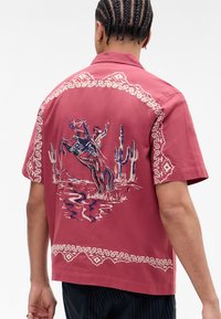 Kortermet skjorte i rose-rød med intrikate hvite broderier langs kraven og kanten, som skildrer en cowboy på en opphisset hest med kaktuser.