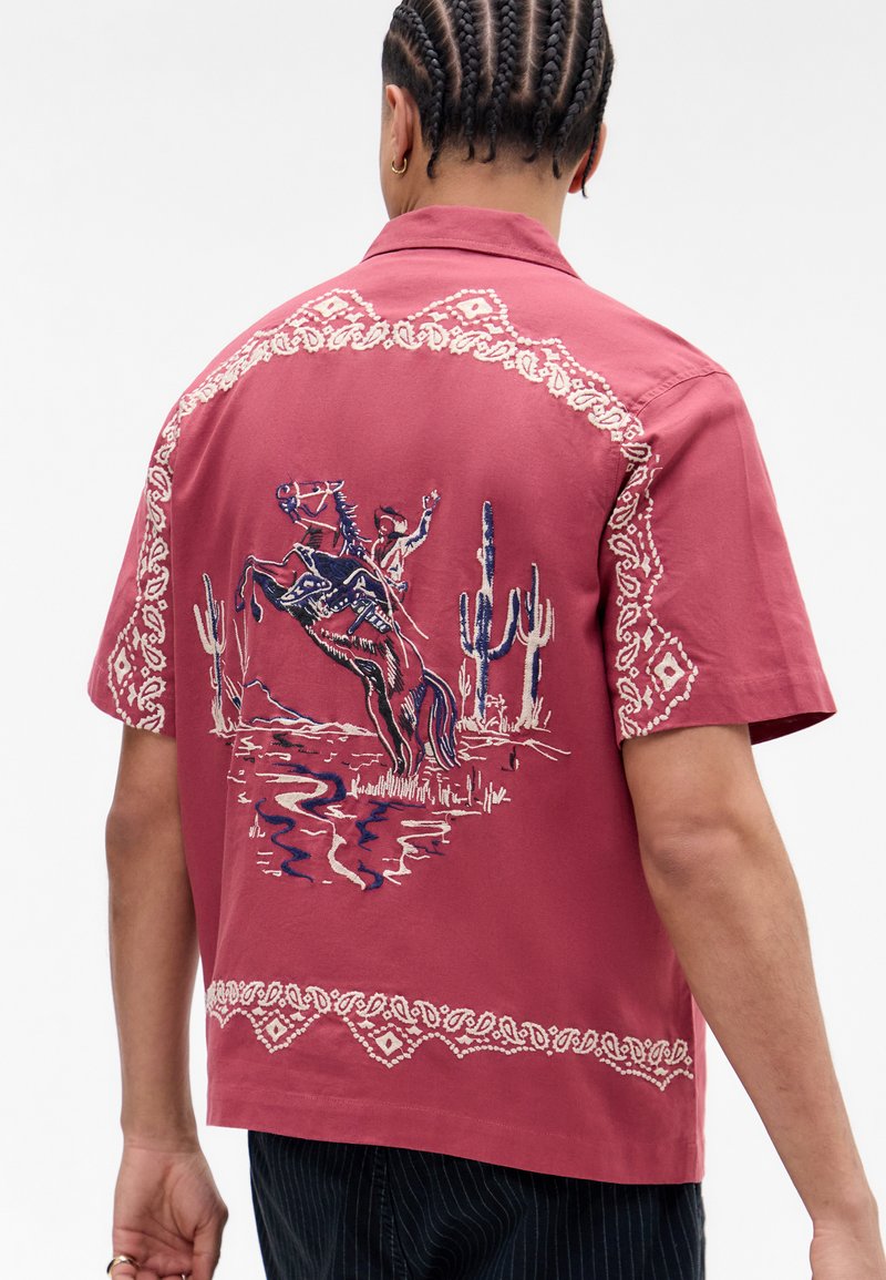 Kortermet skjorte i rose-rød med intrikate hvite broderier langs kraven og kanten, som skildrer en cowboy på en opphisset hest med kaktuser.