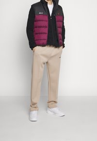 Man i svart långärmad tröja, lila och svart vadderad väst, beige byxor och vita sneakers står med händerna i fickorna.