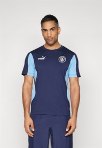 Námořnická modrá sportovní košile se světle modrými bočními panely, zdobená logem Puma na hrudi a emblémem Manchester City. Design s krátkým rukávem.
