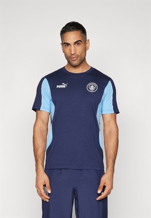 Marineblå sportshem med lyseblå sidepaneler, med Puma-logo på brystet og Manchester City-emblemet. Kortærmet design.
