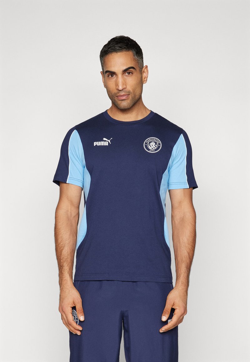 Námořnická modrá sportovní košile se světle modrými bočními panely, zdobená logem Puma na hrudi a emblémem Manchester City. Design s krátkým rukávem.