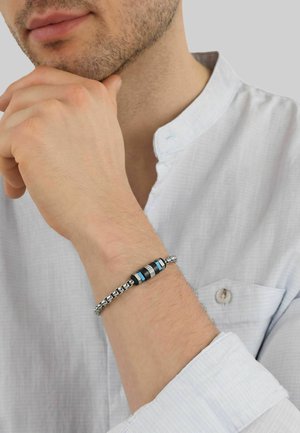 Hombre con camisa blanca apoyando la barbilla en la mano, lleva una pulsera enlazada plateada y negra con detalles azules en la muñeca.