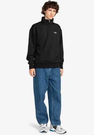 Black sweatshirt with a high collar and half zip. Front features a small white logo. Paired with loose blue denim jeans and sporty sneakers.