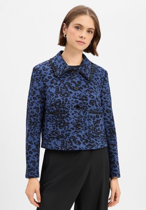 Jeune femme portant une veste bleu et noir à imprimé léopard avec des boutons noirs et deux poches avant, associée à un pantalon noir, debout sur fond blanc.